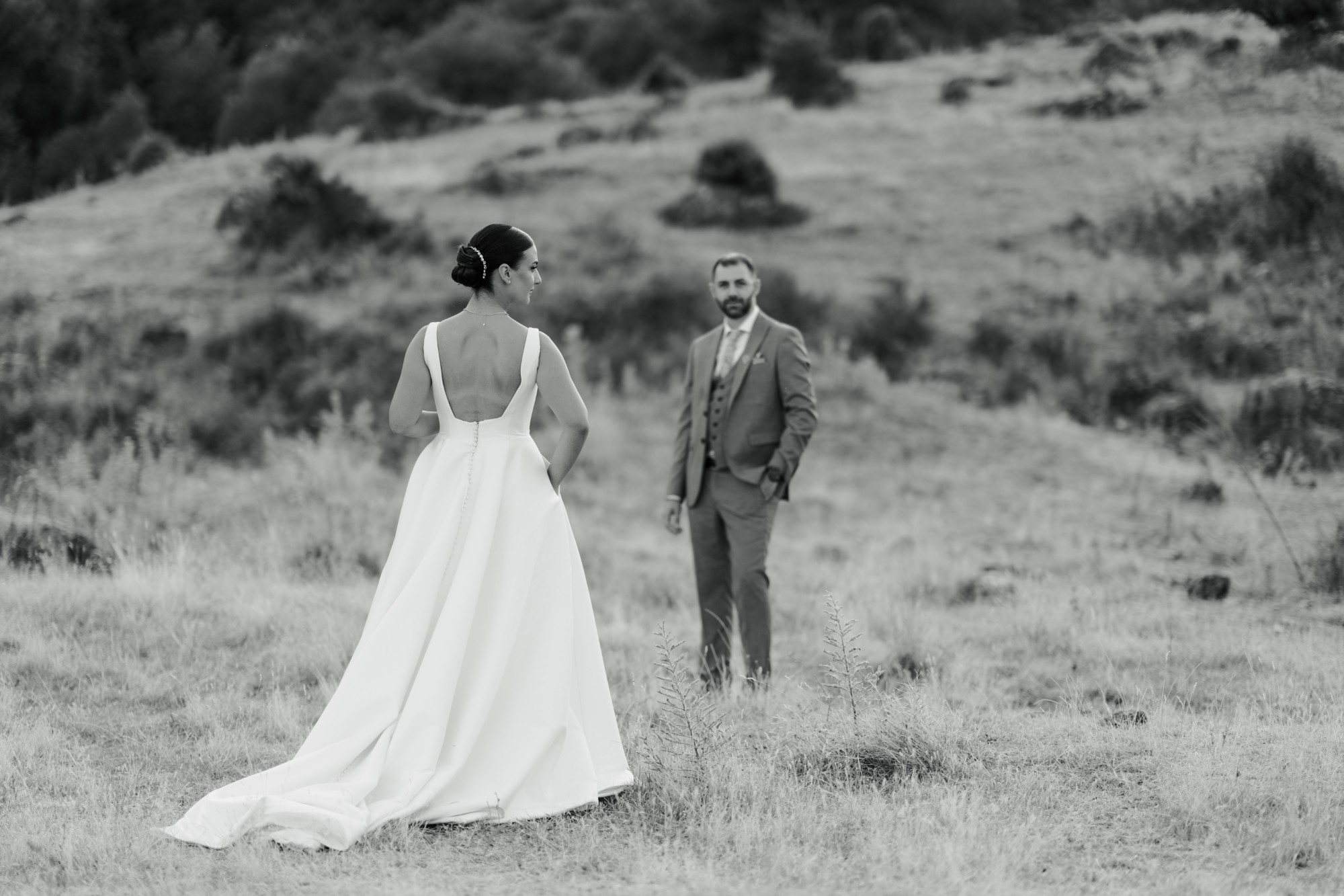 The bride, seen from behind, looks across a meadow at the groom